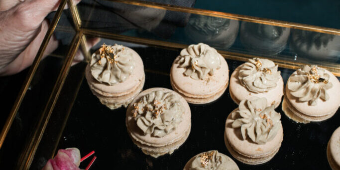 A hand holds black cards with gold "intel | aws" text above a tray of decorated macarons and sliced radish, showcasing elegant party catering.
