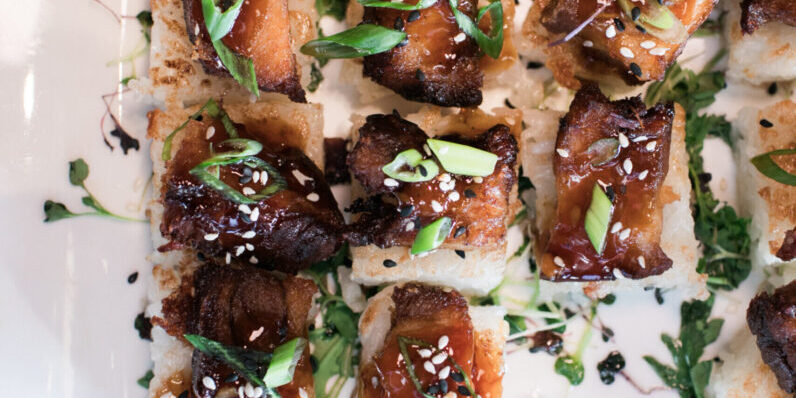 Rows of bite-sized appetizers featuring glazed meat on crispy rice squares, garnished with sesame seeds and sliced green onions, arranged on a white plate—perfect for stylish party catering.