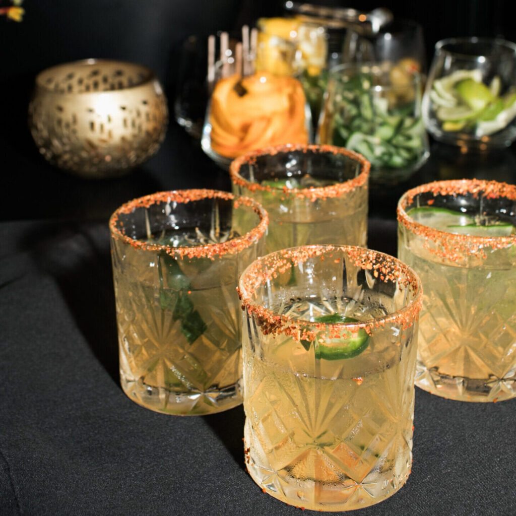 Four cocktails with chili powder rims and lime garnish on a black table, perfect for your next Corporate Catering Seattle event, with assorted garnishes and a candle holder in the background.