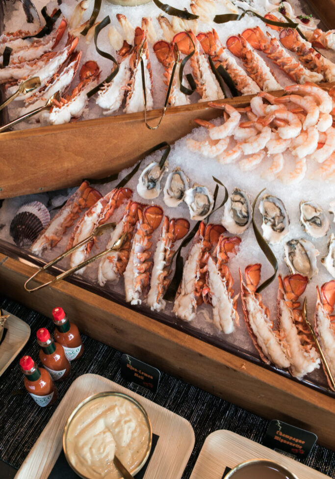 A seafood display perfect for party catering, featuring lobster tails, shrimp, oysters on ice, assorted sauces, and bottles of hot sauce arranged elegantly on a serving table.