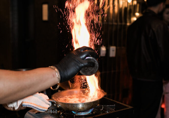 A person wearing a black glove pours liquid into a pan on a portable stove, causing flames to rise, while preparing ingredients for catering for fundraisers.