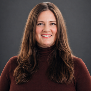 A woman with long brown hair wearing a maroon turtleneck sweater, smiling in front of a dark gray background.