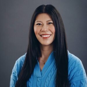 A woman with long black hair, wearing a blue sweater, smiles at the camera against a plain dark background.