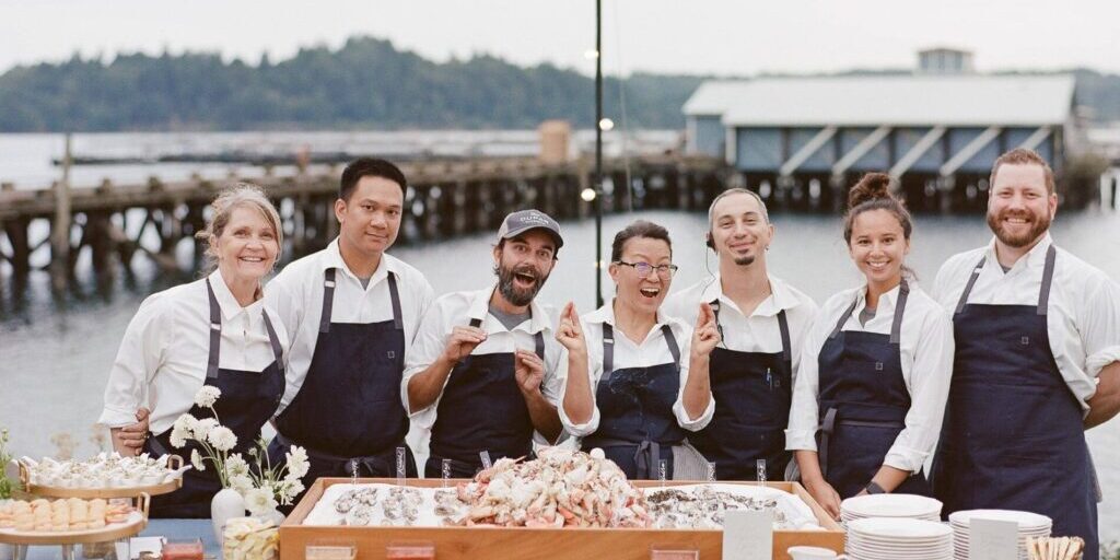 A group of seven smiling people in chef uniforms from About Lisa Dupar Catering Seattle stand behind a seafood display table outdoors near a dock and water.