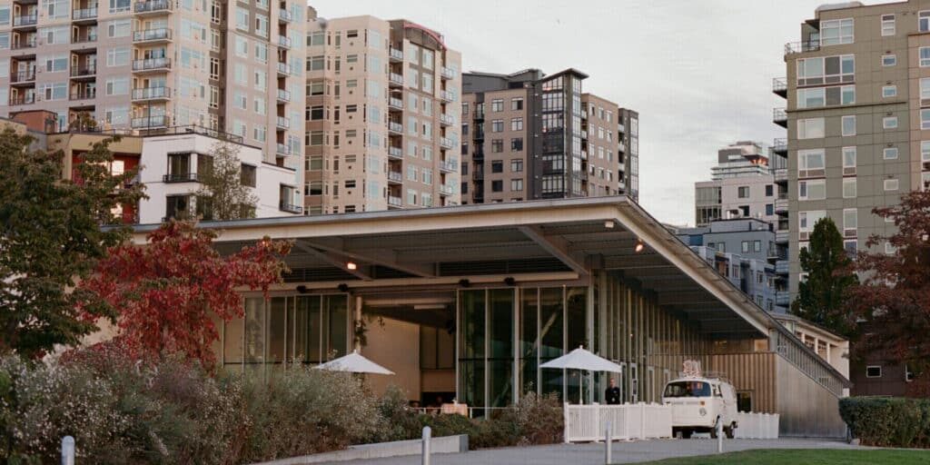 A modern, glass-walled building with a slanted roof stands before tall apartment buildings in an urban area. A white truck, possibly belonging to event vendors in Seattle, is parked beside the building.