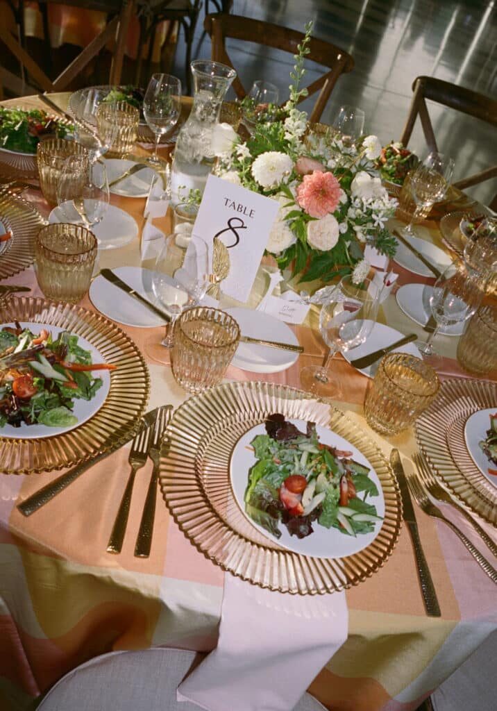 A round table set for a formal event by event vendors in Seattle features gold-rimmed plates, gold utensils, salads, glasses, and a floral centerpiece. A card marked "Table 8" is placed in the middle.