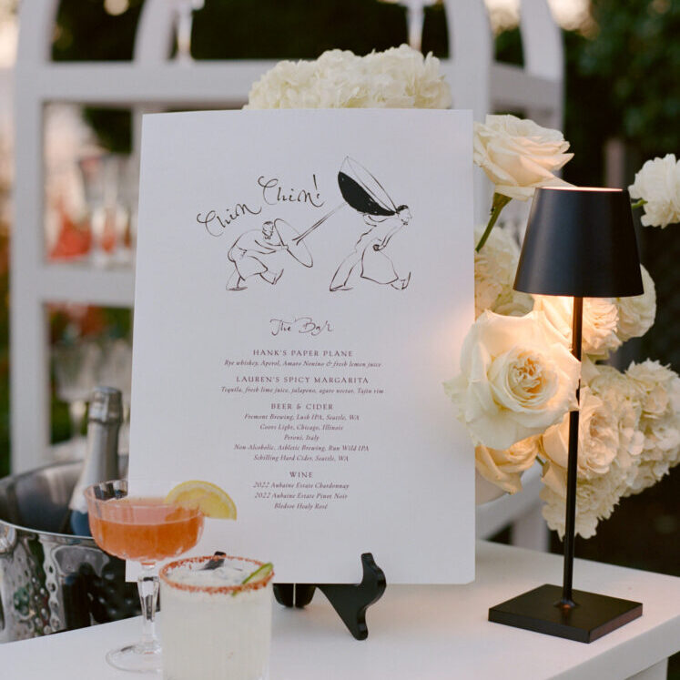 A party catering bar menu is displayed on an easel with two cocktails in front, surrounded by white roses and a small black lamp on a white counter.