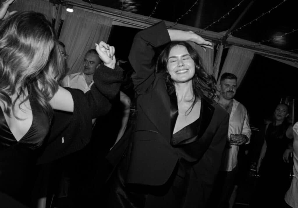 A woman smiles with her eyes closed and dances at a crowded indoor event in Seattle, surrounded by other people in semi-formal attire and event vendors.