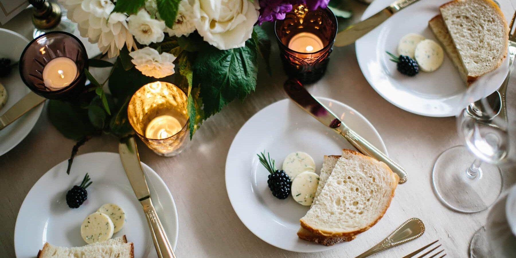 Table setting with plates of bread, butter rounds, and blackberries, surrounded by candles, cutlery, and a floral centerpiece—perfect for elegant wedding and event catering.