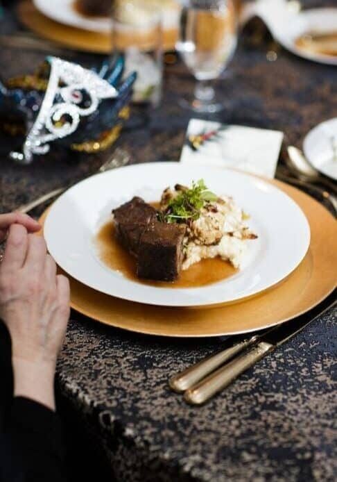 A plate with braised beef on mashed potatoes and sauce, set on a gold charger with silverware, on a patterned tablecloth—ideal catering for fundraisers. A decorative mask and glass complete the elegant background.