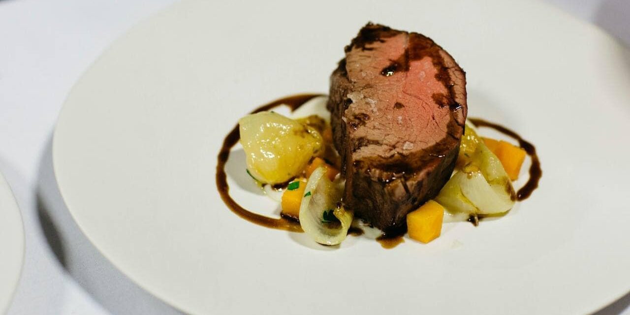 A slice of medium-rare beef tenderloin served with roasted vegetables and a drizzle of sauce on a white plate—an elegant option perfect for catering for fundraisers.
