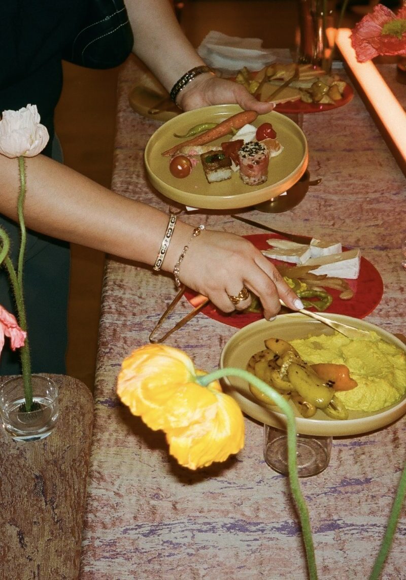 Two people serve food onto plates from a table with various dishes, including vegetables, cheese, dip, and sushi—an inviting spread perfect for corporate catering Seattle—decorated with vases of yellow and orange flowers.