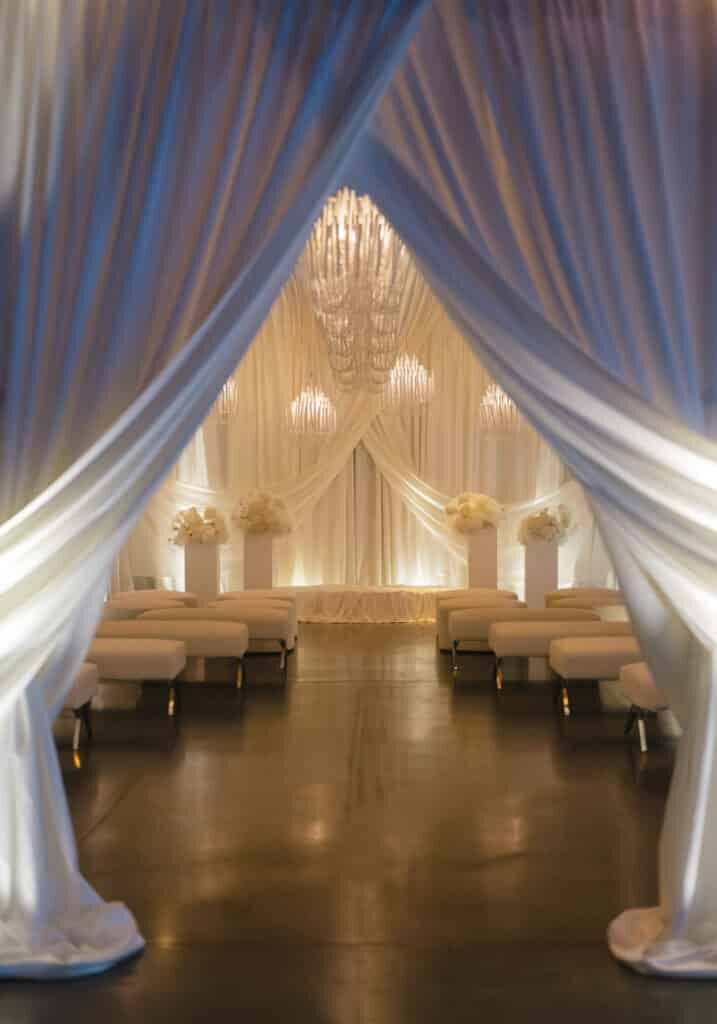 A wedding ceremony space decorated with white draped fabric, chandeliers, and floral arrangements is brought to life by top event vendors in Seattle, with rows of white benches lining either side of the aisle.
