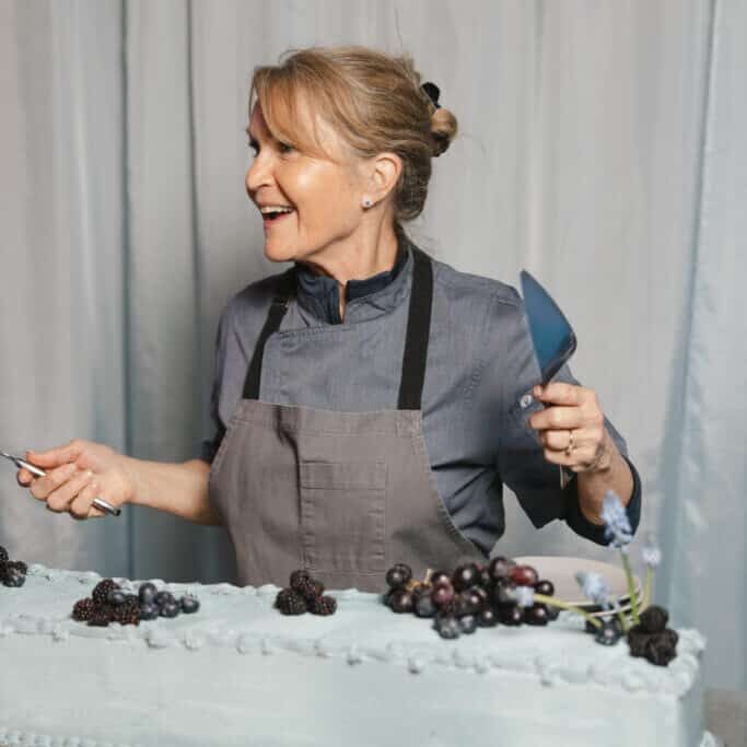 Woman in a gray apron and blue shirt smiles while decorating a large cake with berries, holding a spatula and pen—showcasing her passion for wedding and event catering.