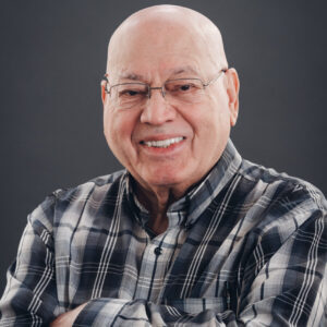 An older man with glasses and a bald head, wearing a plaid shirt, smiles with his arms crossed against a plain dark background.