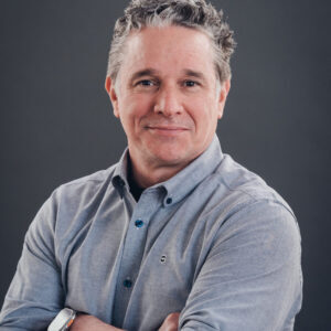 A middle-aged man with short, curly gray hair wearing a light gray button-up shirt stands with arms crossed against a plain dark background.