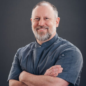 A middle-aged man with a gray beard and short hair, wearing a gray chef’s jacket, stands against a plain dark background with his arms crossed, smiling at the camera.