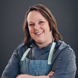 A woman wearing a gray chef’s coat and light blue apron stands with her arms crossed, smiling at the camera against a plain dark background.