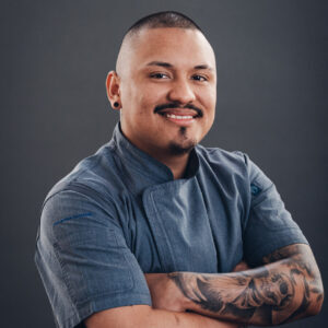 A man with short hair and tattoos on his right arm wears a blue chef's jacket and smiles with his arms crossed in front of a dark gray background.
