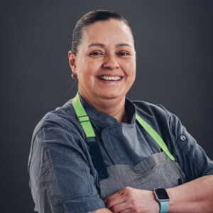 Smiling person wearing a gray chef coat and apron, with arms crossed, against a plain dark background.