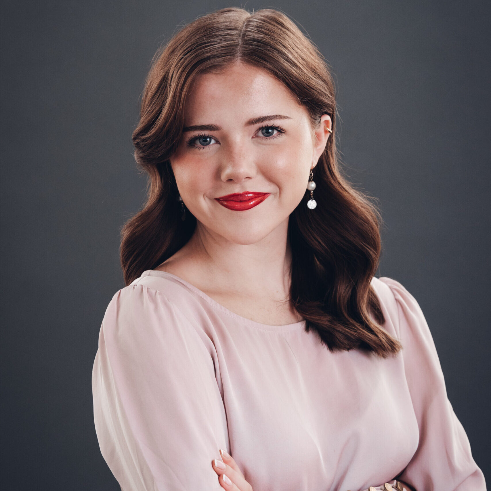 A woman with wavy brown hair, red lipstick, and pearl earrings, wearing a light pink blouse, stands with arms crossed against a dark gray background.