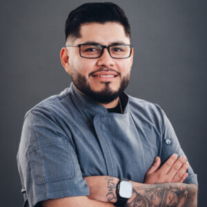 A man with glasses, a beard, and tattoos wearing a gray chef's jacket stands with arms crossed, smiling against a plain gray background.