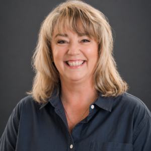 Smiling woman with shoulder-length blonde hair wearing a navy blue button-up shirt, posed against a plain dark background.