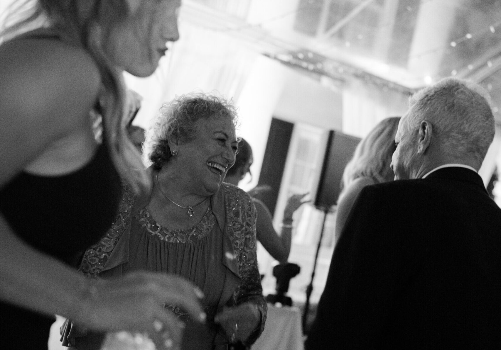A group of people, including an older woman smiling, are socializing and dancing at an indoor event in black and white, highlighting the vibrant atmosphere often seen at gatherings with top Catering Services in Seattle.