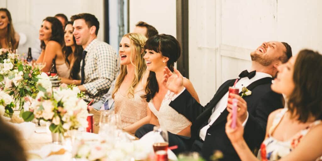 A wedding party sits at a decorated table, smiling and laughing during a celebration. With exquisite wedding catering Seattle, the bride and groom are at the center, surrounded by bridesmaids and groomsmen.
