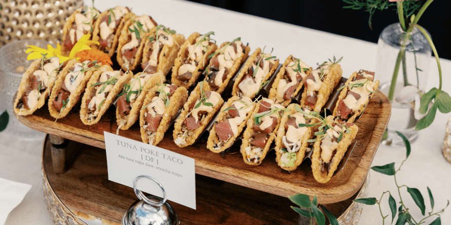 Seattle Catering A wooden stand displaying neatly arranged mini tuna poke tacos, garnished with herbs, on a table set with a single rose in a vase and a small pair of scissors.