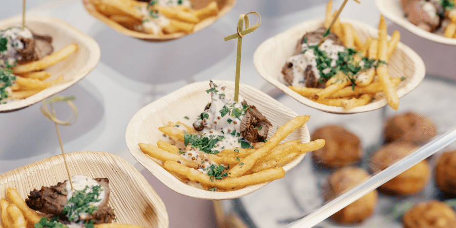 Small bamboo plates with French fries, sliced beef topped with sauce and herbs, each served with a wooden pick; ideal for Corporate Catering Seattle. A tray of bread rolls is visible in the background.