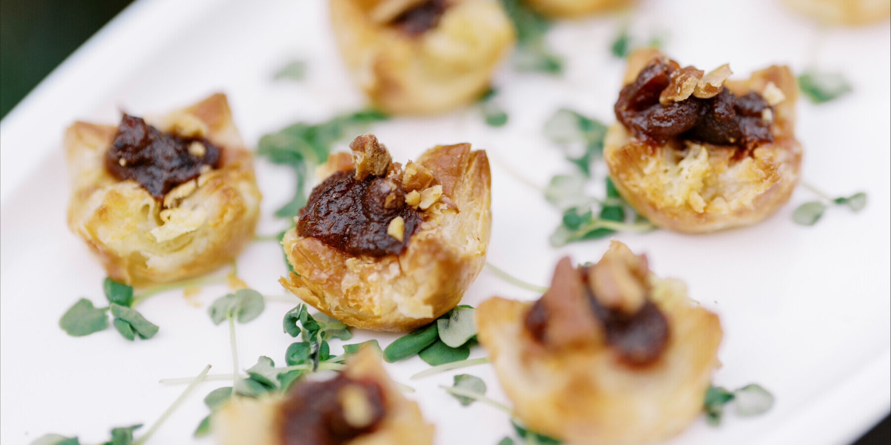 A white platter with several bite-sized puff pastry appetizers, perfect for catering for fundraisers, topped with a dark spread and small garnish, arranged on a bed of microgreens.