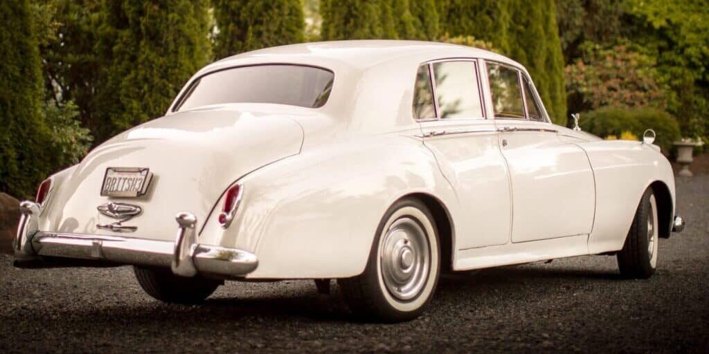 A white vintage luxury car is parked on a gravel driveway, surrounded by greenery and trees—an elegant choice for event vendors in Seattle seeking a timeless backdrop.